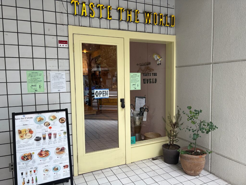 TASTE THE WORLD 新宿店の外観。黄色いドアと店名の看板、店頭にメニューの立て看板
