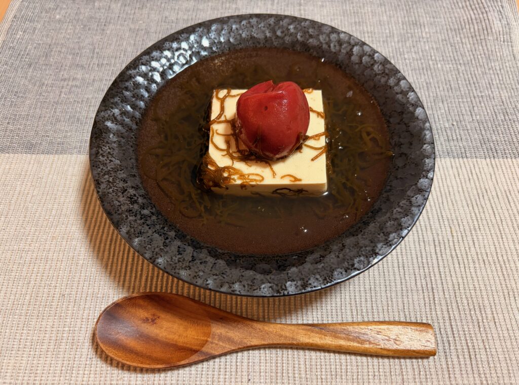 黒い丸皿にのった豆腐ともずくに梅干しを添えた軽食、木のスプーンを添えて