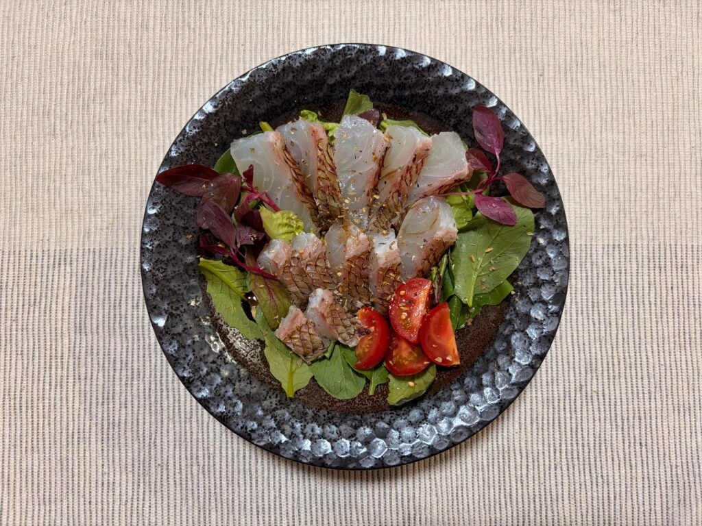黒い器に盛られた鯛の焼き霜造りのカルパッチョ、赤い葉野菜とミニトマト添え